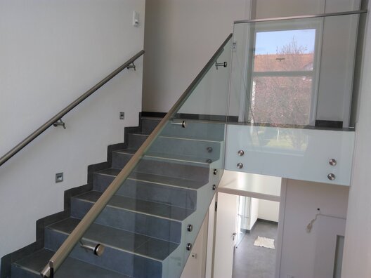 Ein Stiegenhaus mit Geländer aus Glas, Handlauf an Mauer und seitlich bei Glasgeländer. 